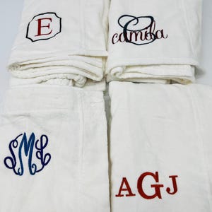 May include: Four white bathrobes with embroidered initials. One robe has a red "E" in a black outlined square. Another has the name "camba" in black script. The third has blue initials "SML" in a decorative font. The last has red initials "AGJ".