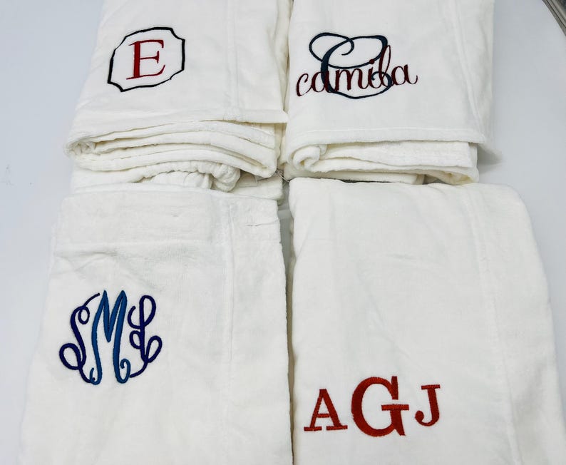 May include: Four white bathrobes with embroidered initials. One robe has a red "E" in a black outlined square. Another has the name "camba" in black script. The third has blue initials "SML" in a decorative font. The last has red initials "AGJ".