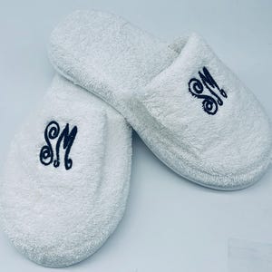 May include: A pair of white terry cloth slippers. Each slipper has the embroidered initials "SM" in a dark blue script. The slippers are fluffy and appear soft, ideal for indoor wear.