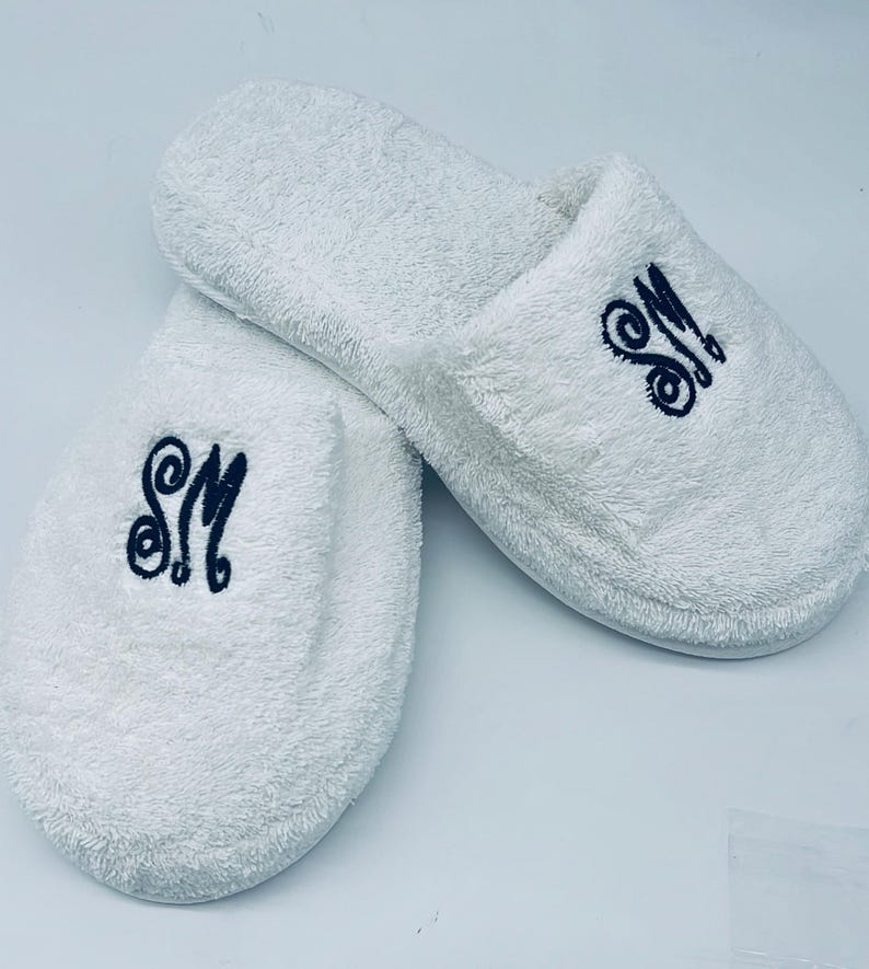 May include: A pair of white terry cloth slippers. Each slipper has the embroidered initials "SM" in a dark blue script. The slippers are fluffy and appear soft, ideal for indoor wear.