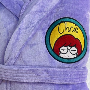 Puede incluir: Batas de ba&ntilde;o de terciopelo morado con un parche bordado circular que presenta una mujer de dibujos animados con cabello rojo y gafas. El parche tiene el texto "Chris'" en un c&iacute;rculo amarillo con un borde turquesa.