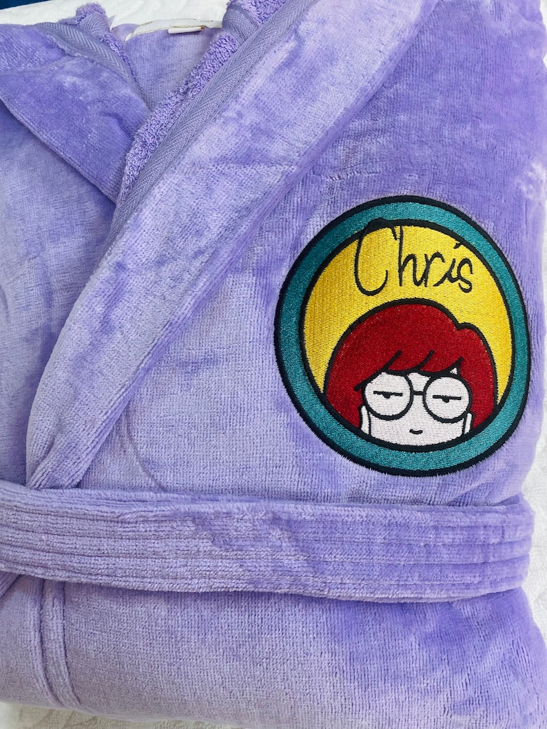 Puede incluir: Batas de ba&ntilde;o de terciopelo morado con un parche bordado circular que presenta una mujer de dibujos animados con cabello rojo y gafas. El parche tiene el texto "Chris'" en un c&iacute;rculo amarillo con un borde turquesa.