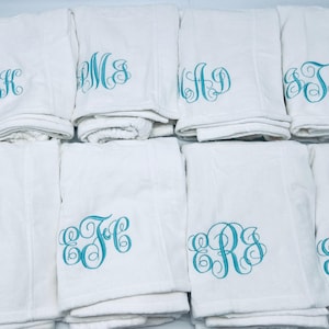 May include: A collection of white towels, each embroidered with a different teal monogram. The towels are folded and stacked, showcasing the personalized initials in a decorative script. The image highlights the custom embroidery.