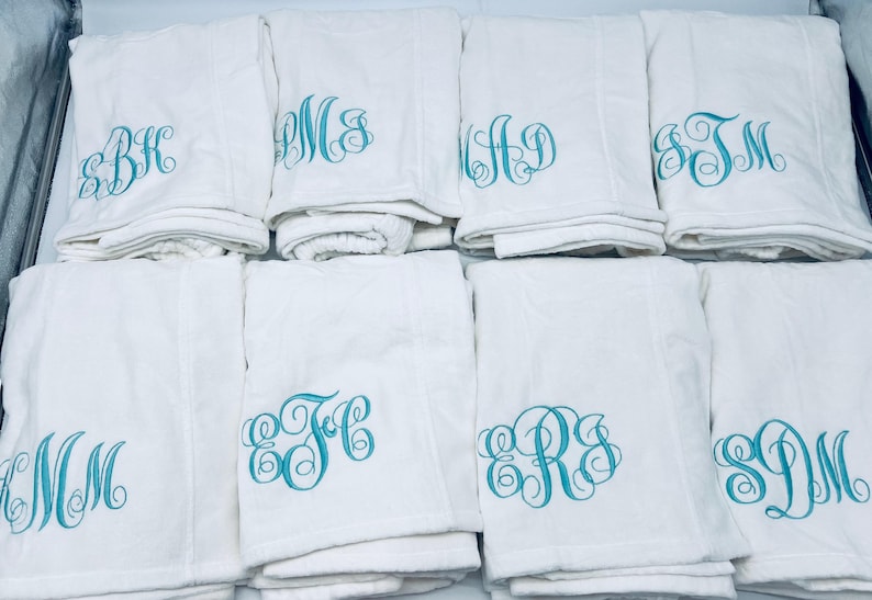 May include: A collection of white towels, each embroidered with a different teal monogram. The towels are folded and stacked, showcasing the personalized initials in a decorative script. The image highlights the custom embroidery.