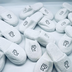 May include: A collection of white terry cloth slippers, each embroidered with the letters "CP" in black. The slippers are arranged on a white surface, showcasing their soft texture and design.