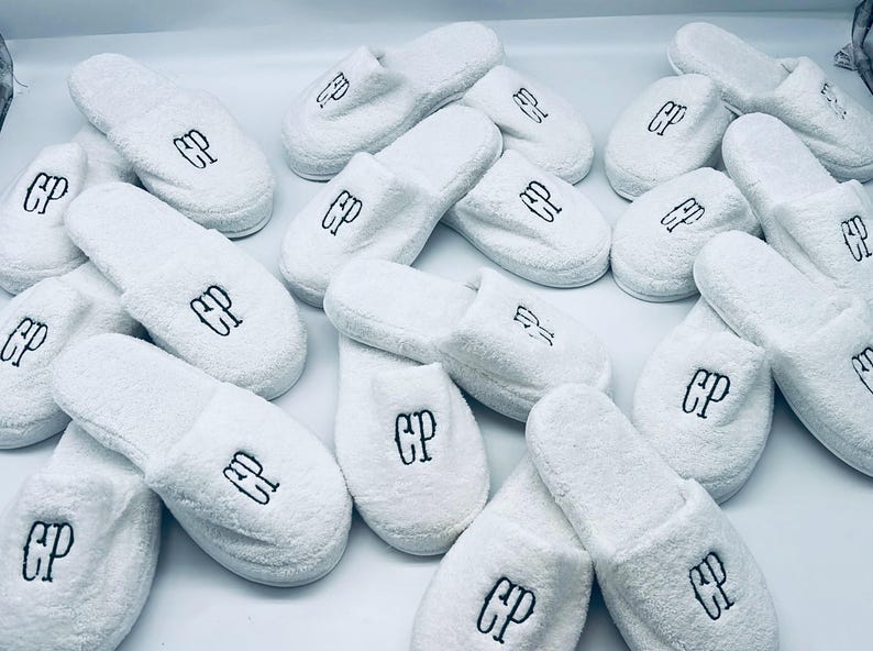 May include: A collection of white terry cloth slippers, each embroidered with the letters "CP" in black. The slippers are arranged on a white surface, showcasing their soft texture and design.