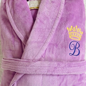 Puede incluir: Una bata de ba&ntilde;o lavanda con una corona amarilla y la letra "B" bordada en la parte delantera. La bata tiene un cintur&oacute;n de atar y un cuello chal.
