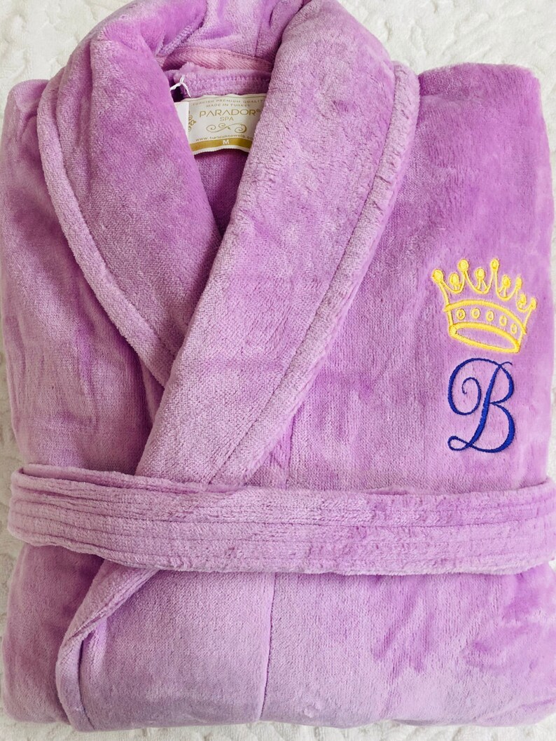 Puede incluir: Una bata de ba&ntilde;o lavanda con una corona amarilla y la letra "B" bordada en la parte delantera. La bata tiene un cintur&oacute;n de atar y un cuello chal.