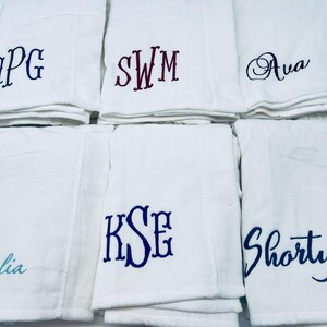 May include: White towels embroidered with various names and initials in blue and burgundy thread. The towels are folded and stacked, showcasing personalized designs for a custom touch. The names include "Natalia" and "Shorty".