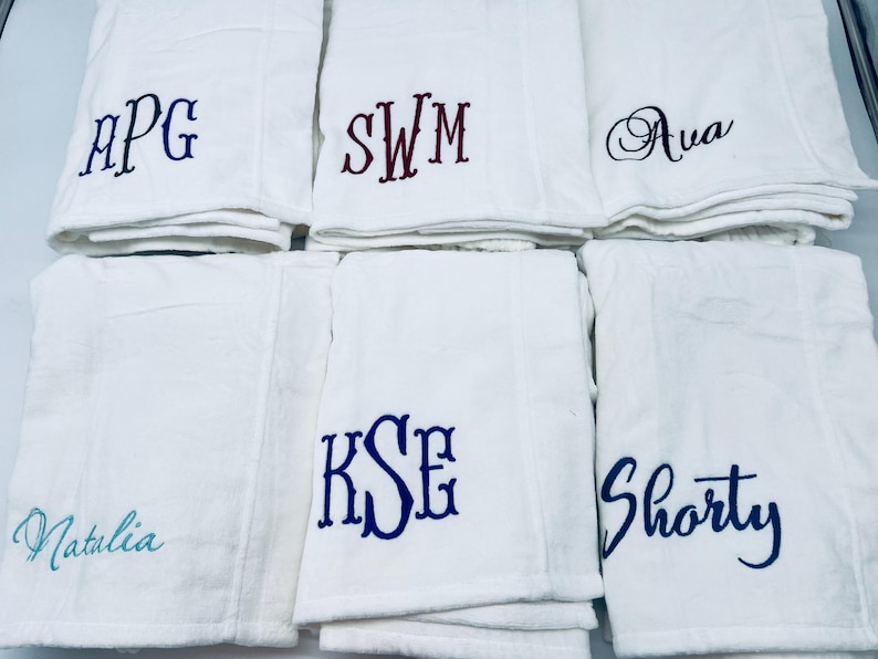 May include: White towels embroidered with various names and initials in blue and burgundy thread. The towels are folded and stacked, showcasing personalized designs for a custom touch. The names include "Natalia" and "Shorty".