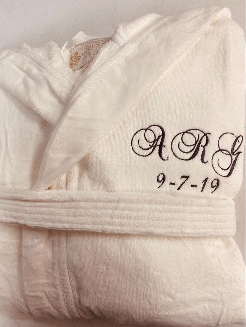 Puede incluir: Batista blanca con capucha con un monograma bordado en negro "ARG" y la fecha "9-7-19".