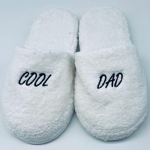 May include: A pair of white terry cloth slippers. The left slipper has the word "COOL" embroidered in navy blue, and the right slipper has "DAD" embroidered in navy blue. The slippers are designed for indoor use.