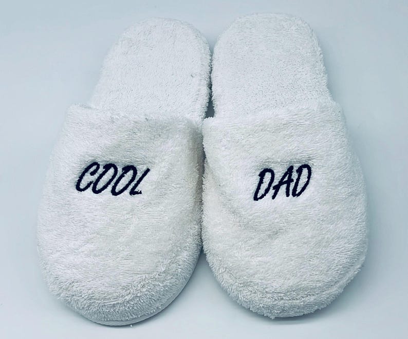 May include: A pair of white terry cloth slippers. The left slipper has the word "COOL" embroidered in navy blue, and the right slipper has "DAD" embroidered in navy blue. The slippers are designed for indoor use.