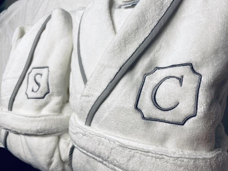 May include: Two white bathrobes with gray piping and embroidered initials. The robe on the left has the letter "S" embroidered on the chest. The robe on the right has the letter "C" embroidered on the chest.
