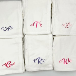 May include: A collection of white terry cloth robes, each embroidered with a unique monogram in shades of pink and purple. The robes are neatly folded and arranged, showcasing the personalized initials. Ideal for spa or bath.