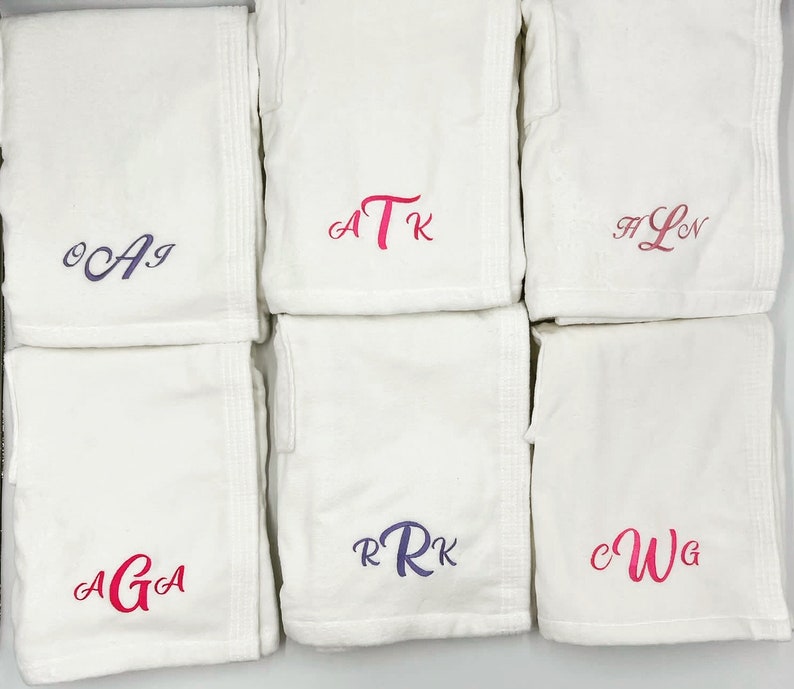 May include: A collection of white terry cloth robes, each embroidered with a unique monogram in shades of pink and purple. The robes are neatly folded and arranged, showcasing the personalized initials. Ideal for spa or bath.