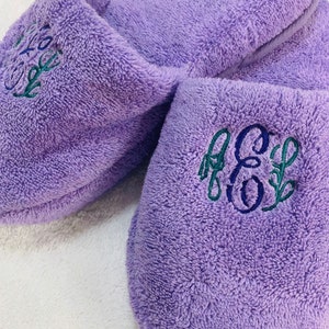 May include: A pair of lavender terry cloth slippers with embroidered initials in blue and green. The slippers have a soft, plush texture and are designed for indoor wear. The embroidery adds a personalized touch.