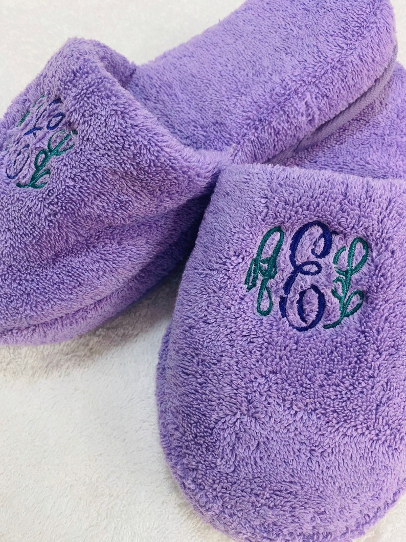 May include: A pair of lavender terry cloth slippers with embroidered initials in blue and green. The slippers have a soft, plush texture and are designed for indoor wear. The embroidery adds a personalized touch.