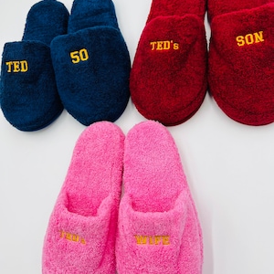 May include: Four pairs of personalized slippers. Two pairs are pink, one pair is blue, and one pair is red. The slippers are embroidered with the words "Wife", "Ted", "50", "Ted's", and "Son".