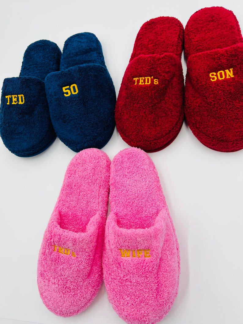May include: Four pairs of personalized slippers. Two pairs are pink, one pair is blue, and one pair is red. The slippers are embroidered with the words "Wife", "Ted", "50", "Ted's", and "Son".