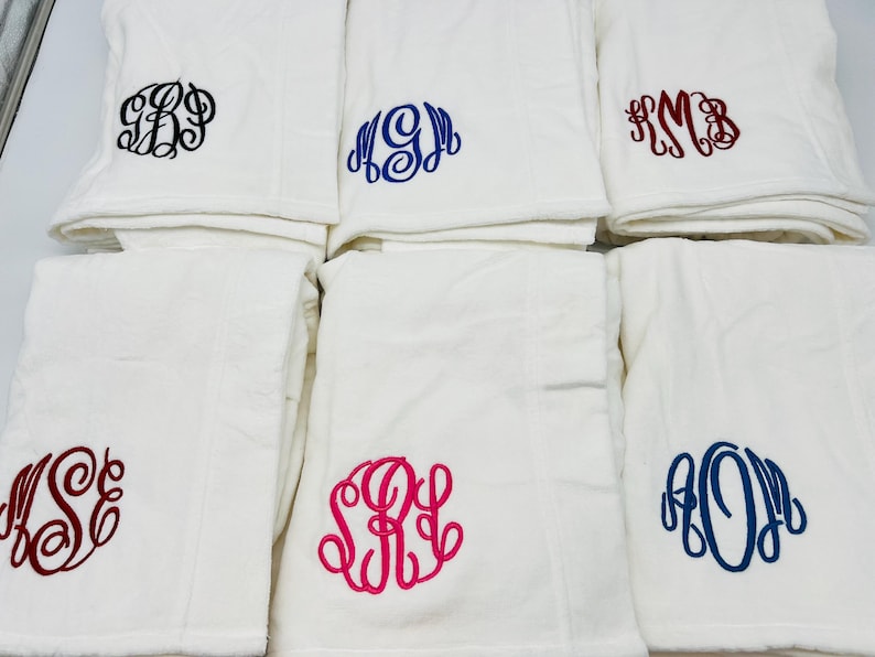 May include: White towels with embroidered monograms in various colors, including black, blue, and red. The towels are folded and stacked, showcasing the personalized initials in a decorative script font. The towels are a soft, plush material.