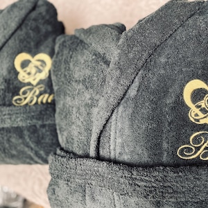May include: Two dark gray bathrobes with gold embroidery. One robe has the word "Bao" embroidered on it, and the other has the word "Boy" embroidered on it. Both robes feature a gold heart with two interlocking rings.