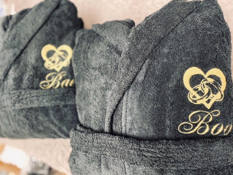 May include: Two dark gray bathrobes with gold embroidery. One robe has the word "Bao" embroidered on it, and the other has the word "Boy" embroidered on it. Both robes feature a gold heart with two interlocking rings.