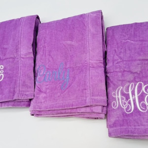 May include: Three folded purple bath towels with white embroidered monograms and a name in blue script. The towels are made of a soft, plush material and are neatly stacked, ready for use.