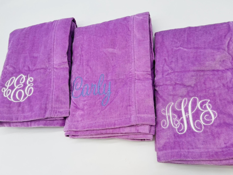 May include: Three folded purple bath towels with white embroidered monograms and a name in blue script. The towels are made of a soft, plush material and are neatly stacked, ready for use.