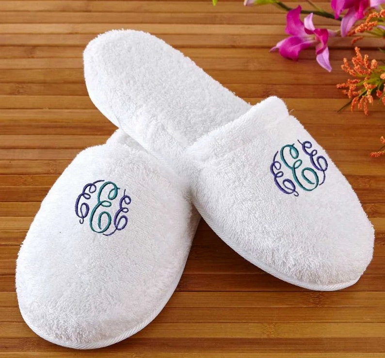 May include: A pair of white terry cloth slippers with an embroidered monogram in blue and teal. The slippers are on a wooden surface, and the design is visible on the top of each slipper. The slippers appear soft and comfortable.