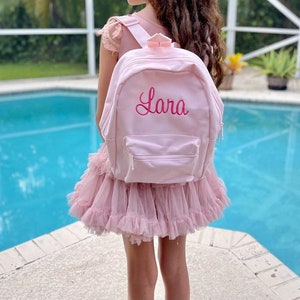 May include: A light pink backpack with the name "Lara" embroidered in a cursive font. The backpack has a front pocket with a zipper and adjustable straps. The background shows a pool and a pink dress.
