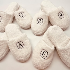 May include: White terry cloth slippers with embroidered initials in black. The slippers are arranged in a group, each displaying a different letter within a black bordered design. The letters include J, R, A, and E.