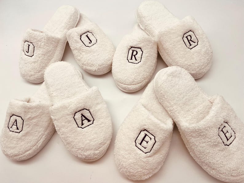 May include: White terry cloth slippers with embroidered initials in black. The slippers are arranged in a group, each displaying a different letter within a black bordered design. The letters include J, R, A, and E.