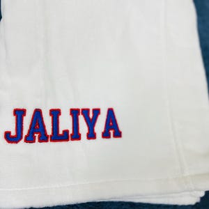 May include: White towel with the name "JALIYA" embroidered in blue letters with a red outline. The towel is folded and placed on a blue surface. The towel appears to be made of a soft, absorbent material.