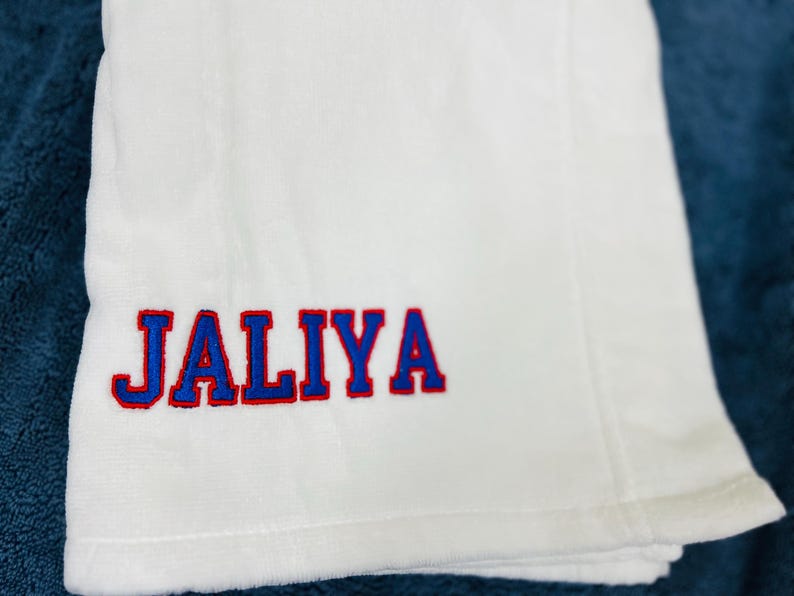 May include: White towel with the name "JALIYA" embroidered in blue letters with a red outline. The towel is folded and placed on a blue surface. The towel appears to be made of a soft, absorbent material.