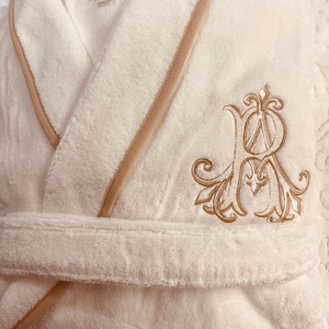 May include: White bathrobe with a beige trim and a gold embroidered monogram. The monogram is a stylized letter "R" with ornate flourishes.