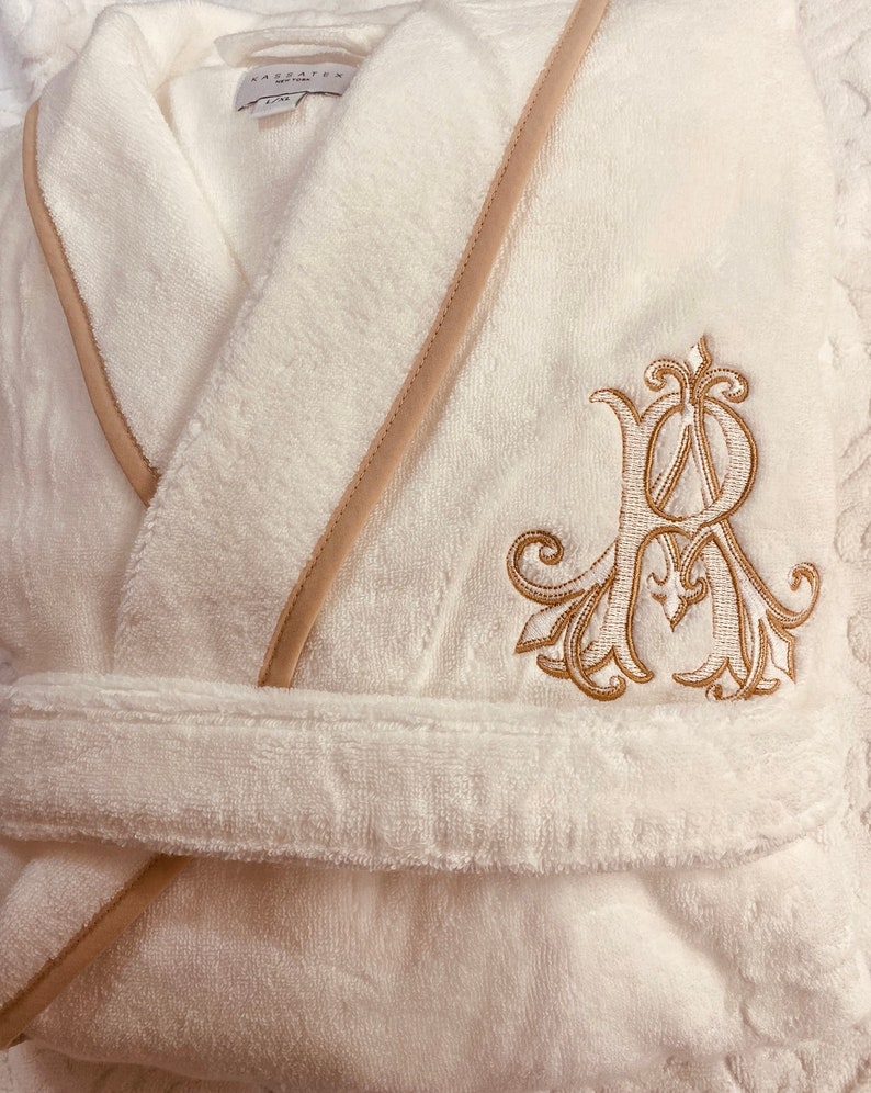 May include: White bathrobe with a beige trim and a gold embroidered monogram. The monogram is a stylized letter "R" with ornate flourishes.