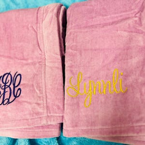 May include: Two pink terry cloth robes with embroidered details. One robe features the initials "JBC" in navy blue, while the other displays the name "Lynlli" in yellow script. The robes are folded and rest on a blue surface.