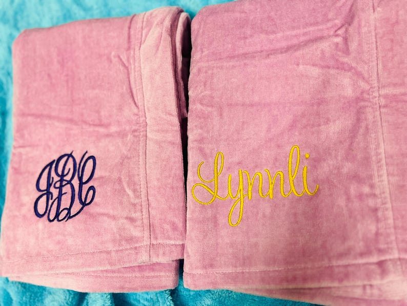 May include: Two pink terry cloth robes with embroidered details. One robe features the initials "JBC" in navy blue, while the other displays the name "Lynlli" in yellow script. The robes are folded and rest on a blue surface.