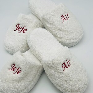 May include: Four white terry cloth slippers. Two slippers have the word "Air" embroidered in red, and the other two have the word "Jefe" embroidered in red. The slippers are arranged on a white background.