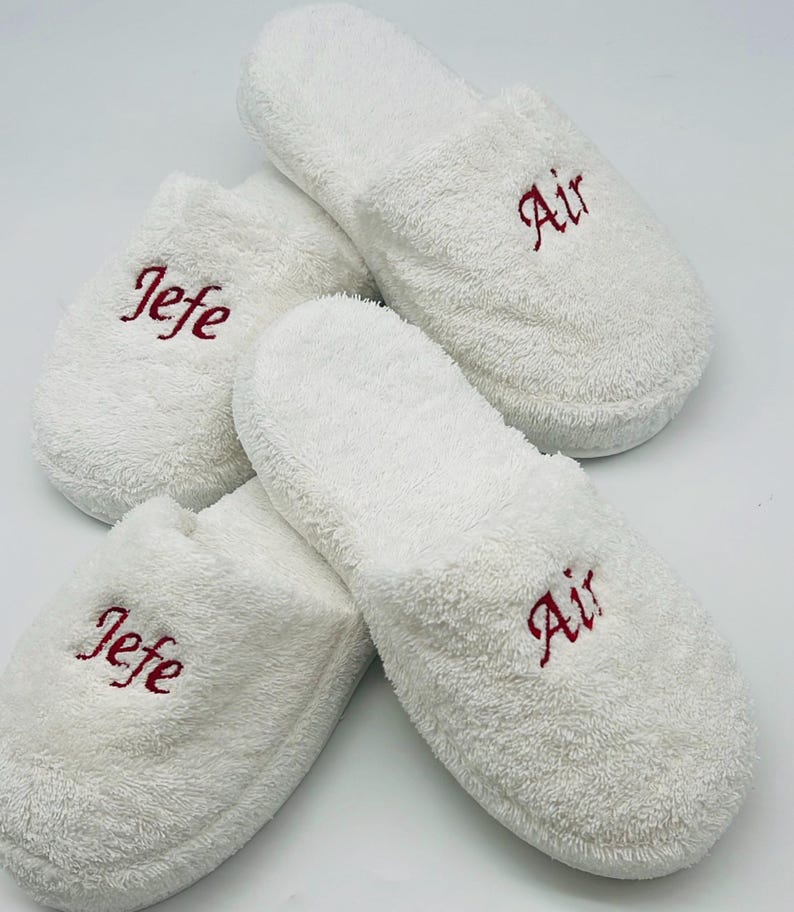 May include: Four white terry cloth slippers. Two slippers have the word "Air" embroidered in red, and the other two have the word "Jefe" embroidered in red. The slippers are arranged on a white background.