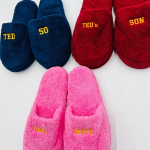 May include: A collection of plush slippers in various colors. Navy blue slippers with "TED" and "50" embroidered in yellow. Red slippers with "TED'S" and "SON" in yellow. Pink slippers with "TED'S" and "WIFE" in yellow.