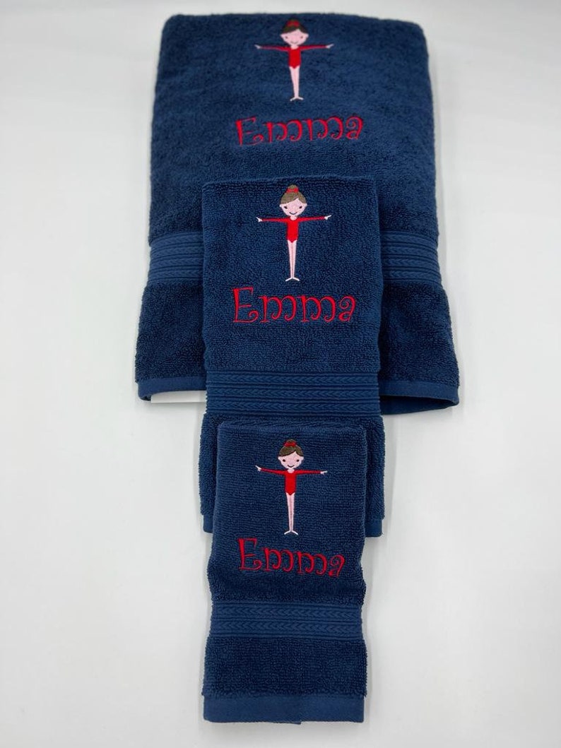 May include: A set of three navy blue towels with embroidered gymnast figures and the name "Emma" in red thread.