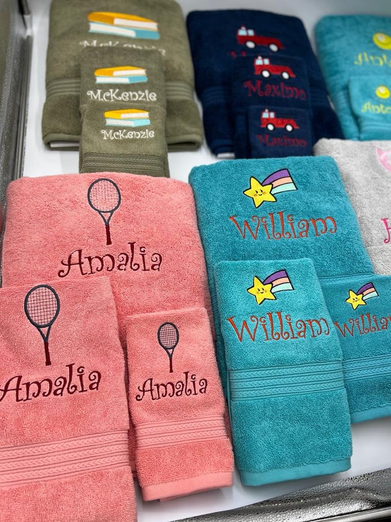 May include: A set of personalized towels with embroidered names and designs. The towels are in various colors, including pink, blue, and green. The designs include a tennis racket, a star with a rainbow, and books.