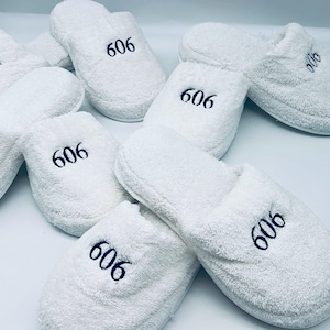 May include: A collection of white terry cloth slippers, each embroidered with the number "606" in navy blue. The slippers are soft and fluffy, perfect for indoor use. The image shows several pairs of these comfortable, spa-like slippers.