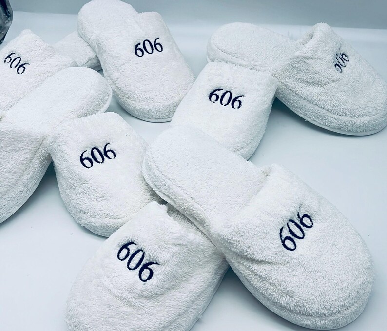 May include: A collection of white terry cloth slippers, each embroidered with the number "606" in navy blue. The slippers are soft and fluffy, perfect for indoor use. The image shows several pairs of these comfortable, spa-like slippers.
