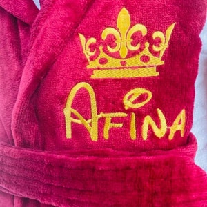 Puede incluir: Bata de color burdeos con una corona bordada en oro y el nombre "AFINA" en la parte delantera.