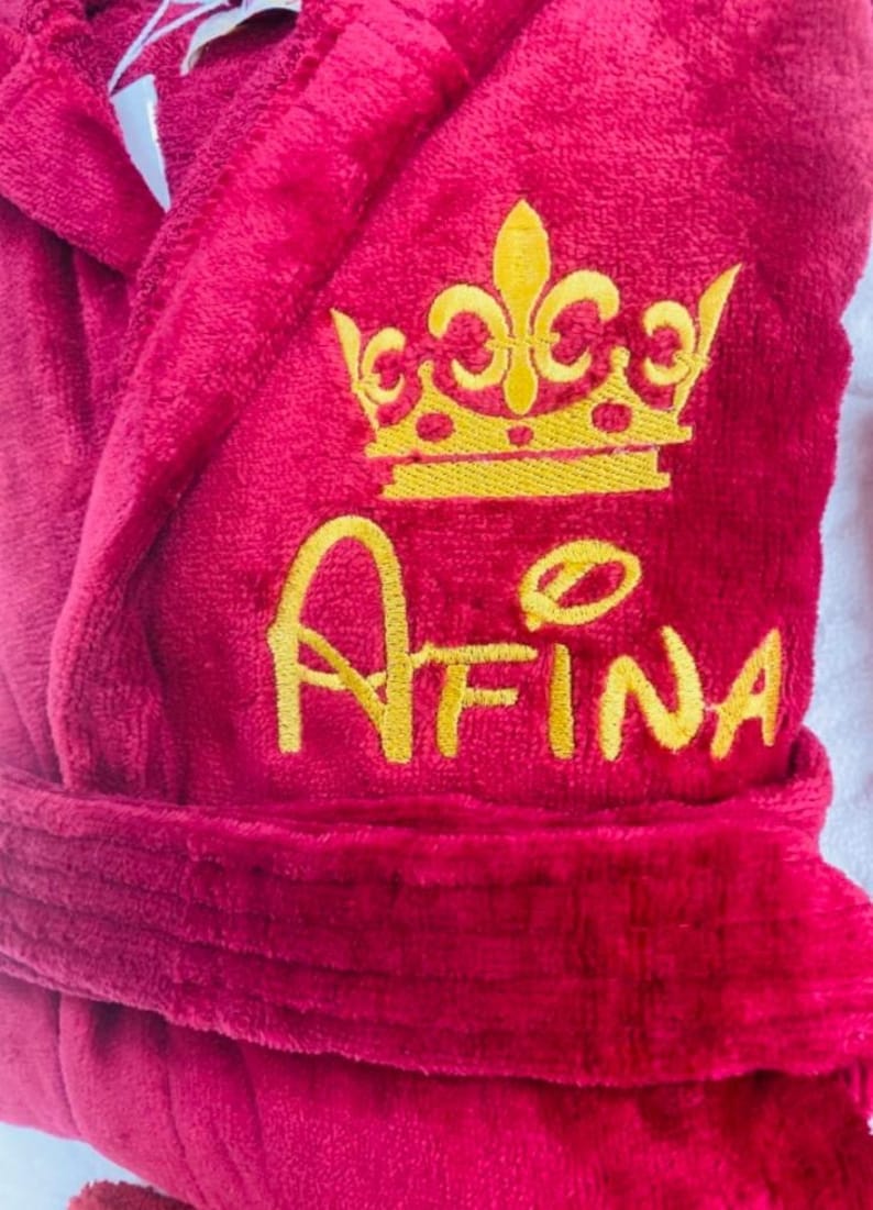 Puede incluir: Bata de color burdeos con una corona bordada en oro y el nombre "AFINA" en la parte delantera.