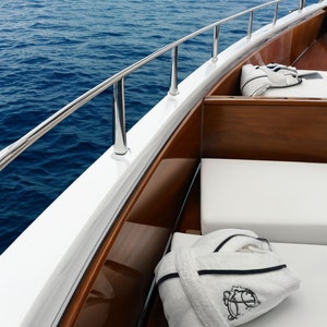 May include: A white bathrobe with black trim lies on a white cushioned seat on a wooden boat deck. The boat is docked at a marina with blue water in the background.