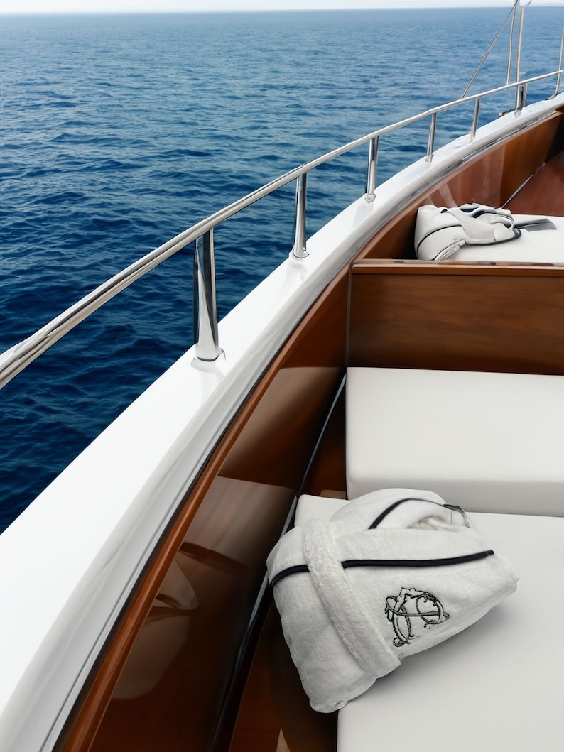 May include: A white bathrobe with black trim lies on a white cushioned seat on a wooden boat deck. The boat is docked at a marina with blue water in the background.